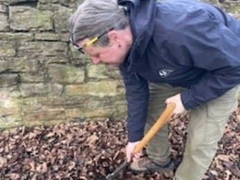 man digging soil covered in leaves with stone wall behind him