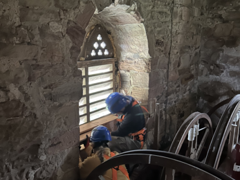 Two workers in safety gear installing bird boxes by a small arched window in a stone-walled room with bell-ringing equipment.
