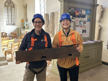 Two people in safety gear hold a wooden bird boxes inside a church with stone columns and arched windows.