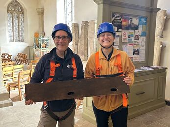 Two people in safety gear hold a wooden bird box for swifts inside a historic church, with stone artifacts and display boards behind them.