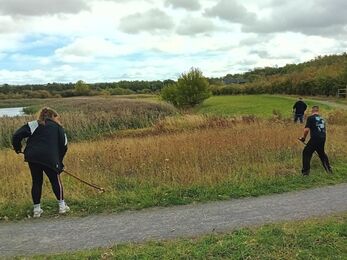Volunteers using hand tools to cut tall grass near a path and water, under a cloudy sky.