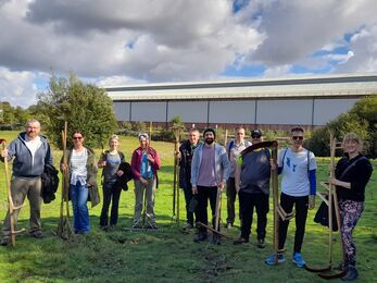 Group of people stand on a grassy field holding traditional farming tools, with an industrial building and trees in the background under a partly cloudy sky.