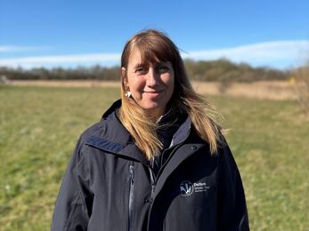 A person wearing a dark Durham Wildlife Trust jacket stands in a grassy field under a clear blue sky, with trees and shrubs in the distance.