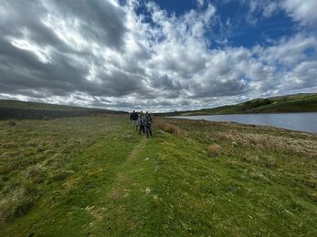 A small group of walkers follow a grassy path beside Grassholme Reservoir under a dramatic sky of thick, rolling clouds. The water glints to the right, while green hills rise in the distance, creating a peaceful scene of volunteers exploring Teesdale’s open countryside.