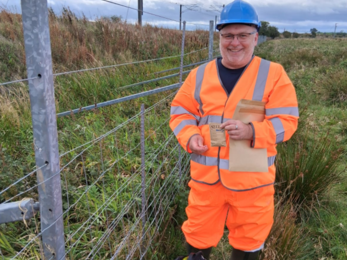 A man wearing a bright orange high‑visibility suit, blue hard hat, and green boots stands beside a metal wire fence in a grassy field. He smiles while holding two brown paper envelopes labeled “SEEDS.” Behind him, railway power lines run parallel to the fence under a cloudy sky. The scene suggests fieldwork or seed collection near railway land.