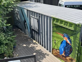 A long metal container painted with a colourful wildlife mural at Rainton Meadows Nature Reserve. The artwork shows a vivid blue‑and‑orange kingfisher perched on a branch against a green leafy background, while a black‑and‑white bird in flight decorates the grey and blue side panels. The container sits on a wooden deck surrounded by greenery, blending art and nature in a creative outdoor space.
