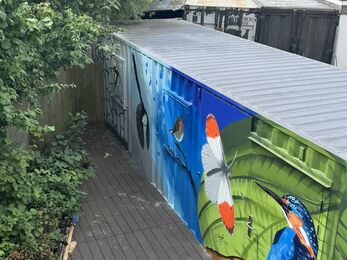 A long metal container painted with a vibrant wildlife mural. The artwork features a black‑and‑white bird in flight on the left, a robin perched on a branch in the centre, and a large butterfly with white wings and orange tips near a bright blue‑and‑orange kingfisher on the right. The backgrounds shift from grey to blue to green leaf patterns, creating a lively nature scene. The container sits outdoors beside greenery and parked vans.