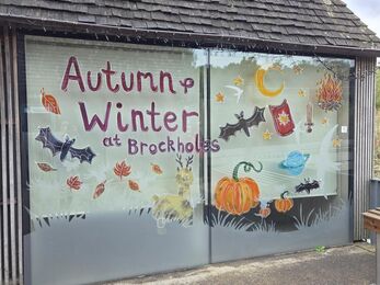 A large glass window decorated with colourful hand‑painted illustrations and the words “Autumn & Winter at Brockholes.” Surrounding the text are bats, pumpkins, falling leaves, stars, a crescent moon, a deer, a campfire, a sword, and a planet with rings. The artwork is cheerful and seasonal, celebrating autumn and winter at the Brockholes nature reserve.