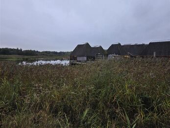A group of thatched‑roof cabins sits beside a calm lake under an overcast sky. Tall reeds and grasses fill the foreground, framing the water and rustic buildings. String lights add a faint warm glow along the huts, creating a tranquil, natural scene.