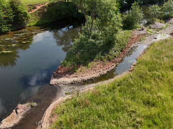 Aerial view of a river bend bordered by grassy banks and trees, with a narrow gravel path following the shoreline. The water curves around a small vegetated peninsula