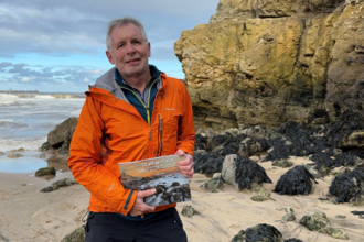Man standing on beach in front of rock formation holding book