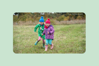 two children wearing winter kit and wellies running across field towards camera