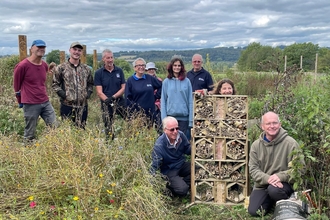 Group of people standing and kneeling around a large bug hotel structure in a field