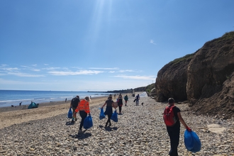 A group of volunteers walk along a beach during a cleanup event, each carrying large blue rubbish bags. The beach is flanked by cliffs on one side and the sea on the other, with a few distant figures and a tent near the shoreline. The sky is clear and sunny.