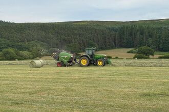 A green tractor pulls a round baler across a grassy field, producing cylindrical hay bales. One bale lies near the baler, with more scattered in the distance. Forested hills surround the rural scene.