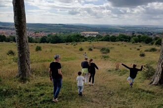 A group of adults and children walk down a grassy hill toward a distant village, surrounded by countryside.