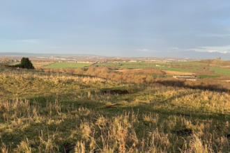 A wide view from a grassy hill overlooking patchwork fields and a distant town under a pale, lightly clouded sky.