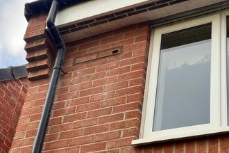 Close-up of a red-brick house wall with a white-framed window, a black downpipe, and a small vent near the roofline beneath a white fascia and black gutter.