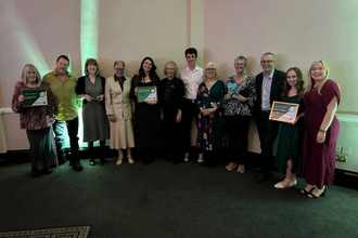 A group of thirteen people stand in a row indoors at an awards event, several holding framed certificates for categories including Community Group, Young Volunteer, and Arts and Culture, with green lighting on the wall behind them.