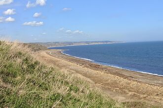 A wide coastal view showing a long, curving beach bordered by grassy cliffs. The shoreline is a mix of sand, pebbles, and scattered driftwood, with calm blue water stretching to the horizon under a mostly clear sky. Distant headlands and cliffs continue along the coast.