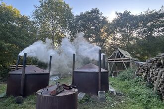 Three large metal charcoal kilns stand in a clearing, each releasing thick white smoke from a central chimney. They sit among dense green woodland, with a wooden shelter and a neatly stacked pile of cut logs to the right.
