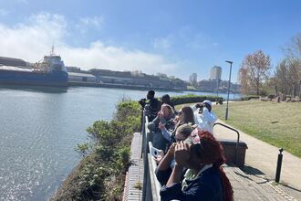 A group of people stand along a riverside walkway on a sunny day, looking out over the water. Some use binoculars or cameras, while others shade their eyes with their hands. A large blue cargo ship is docked across the river, and tall buildings rise in the background under a clear blue sky. The scene includes greenery, a brick wall, and a paved path beside the river, capturing a peaceful moment of observation and community outdoors.