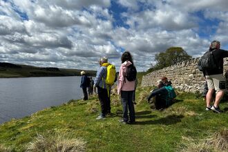    A group of volunteers stand and sit along the grassy edge of Grassholme Reservoir under a dramatic sky filled with scattered clouds. They wear outdoor gear and backpacks, looking out over the water and surrounding hills, with a stone wall running beside them — capturing a peaceful moment of wildlife observation in Teesdale’s open countryside.