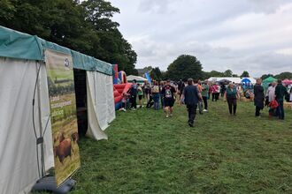 People walk through a grassy field at an outdoor fair with tents and stalls offering land management advice and other displays. A banner showing grazing cattle hangs on a white and green tent to the left, while visitors mingle near colourful booths and hay bales under an overcast sky.