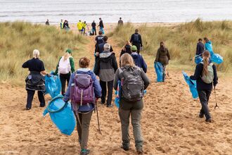 A group of volunteers walk down a sandy path between grassy dunes toward the beach, carrying blue litter bags and pickers for a cleanup. The sea stretches out ahead under a cloudy sky, and participants wear outdoor clothing and backpacks, ready to collect rubbish along the shore.
