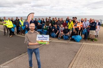    A large group of volunteers stand together near the seafront after a beach clean, smiling and holding blue litter bags and pickers. In the foreground, a person holds a sign reading “Big Autumn Clean #CleanerDurham.” Behind him, participants in high‑visibility jackets and casual outdoor clothing pose with the sea and cloudy sky in the background, celebrating their collective cleanup effort.