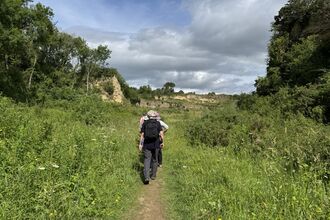    A group of hikers walks along a narrow dirt path surrounded by tall green grass and wildflowers. Steep, rocky cliffs rise on either side, covered in dense vegetation. The sky above is partly cloudy, with patches of blue breaking through. The scene captures a peaceful outdoor walk through a lush, natural landscape.