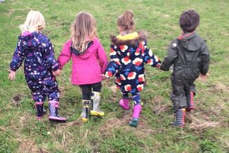 Four young children in colourful outfits and wellies holding hands and walking away from the camera