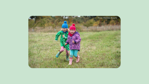 two children wearing winter kit and wellies running across field towards camera