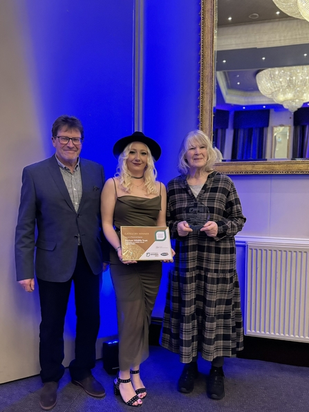 Three people in front of mirror with middle person holding award. 