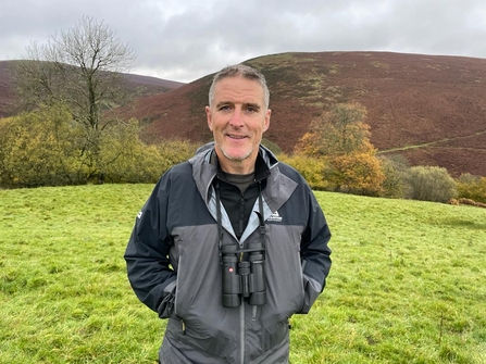 Iolo Williams smiling at the camera. Hills in the background.