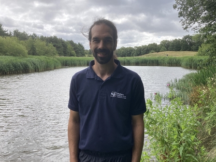 Man standing in front of lake with fields and trees across water in background