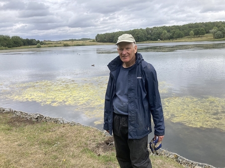 Man standing in front of lake with fields and trees across water in background