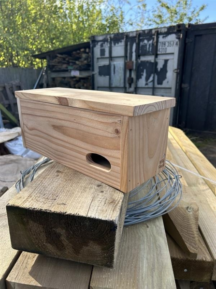 Wooden bird house with oval entrance sits on top of stacked planks and wire in outdoor workshop.