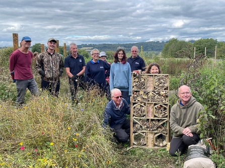 Group of people standing and kneeling around a large bug hotel structure in a field