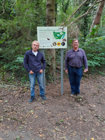 Two people standing next to an interpretation sign in front of woodland area