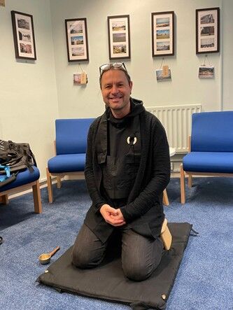 A person dressed in black kneels on a black meditation cushion in a room with blue carpet and blue chairs. They are smiling at the camera. Behind them is a white radiator and a wall with two rows of framed photographs. A small bowl with a handle sits on the floor beside the cushion. The setting appears calm and informal, possibly used for meditation or relaxation.