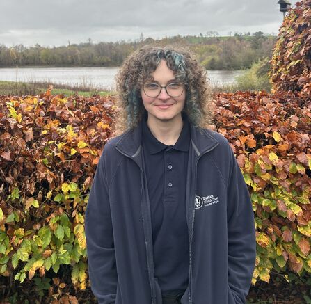 Lyze Bell wearing Durham Wildlife Trust branded clothing standing in front of autumnal hedge with lake in background