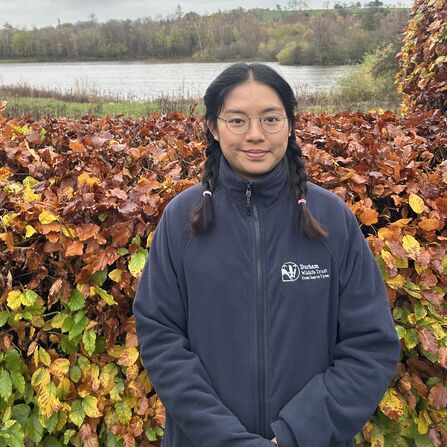 Wai Kan wearing Durham Wildlife Trust branded clothing standing in front of autumnal hedge with lake in background