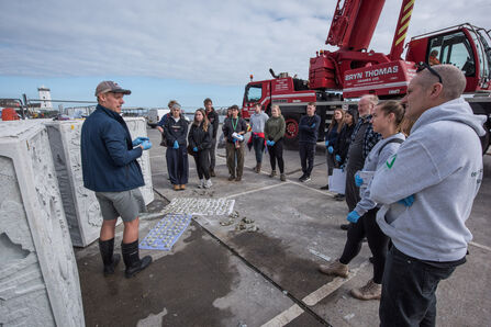 Outdoor marine science workshop at a dock, with speaker and group examining samples near a crane.