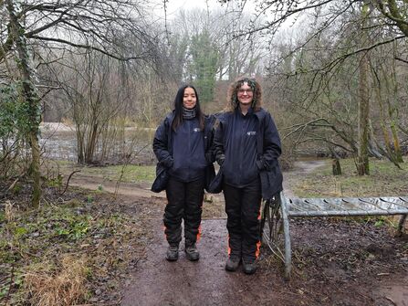 Two conservation workers in dark outdoor gear stand on a muddy riverside path in winter woodland, with a metal bench beside them.