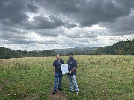 Two people stand in an open field under dark storm clouds, holding a large printed poster between them.