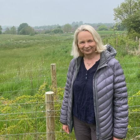  Person standing by a wire fence in a green field, wearing a grey puffer jacket under an overcast sky.