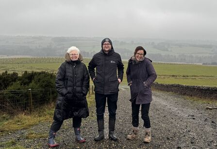 Three people in winter coats and hats stand on a gravel path in open countryside, with rolling fields and cloudy skies behind them.