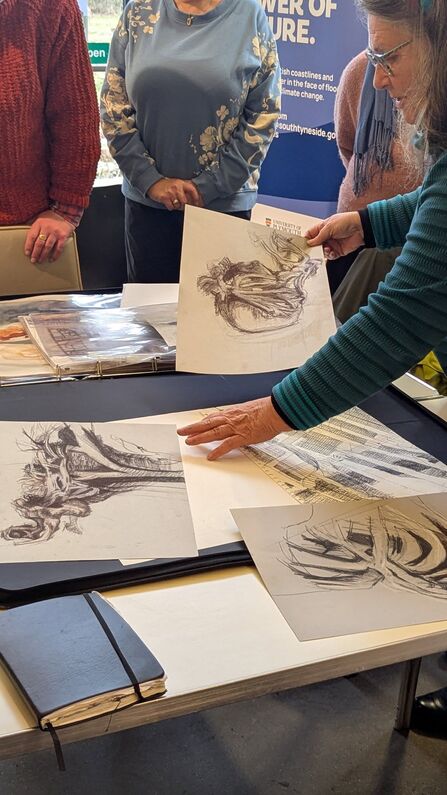 A group of people stand around a table covered with detailed black‑and‑white drawings. One person holds up a sketch while others look on. A banner in the background reads “Power of Nature” with logos for South Tyneside Council and the University of Plymouth.