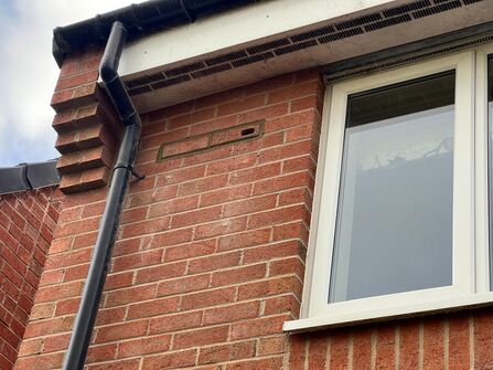 Close-up of a red-brick house wall with a white-framed window, a black downpipe, and a small vent near the roofline beneath a white fascia and black gutter.